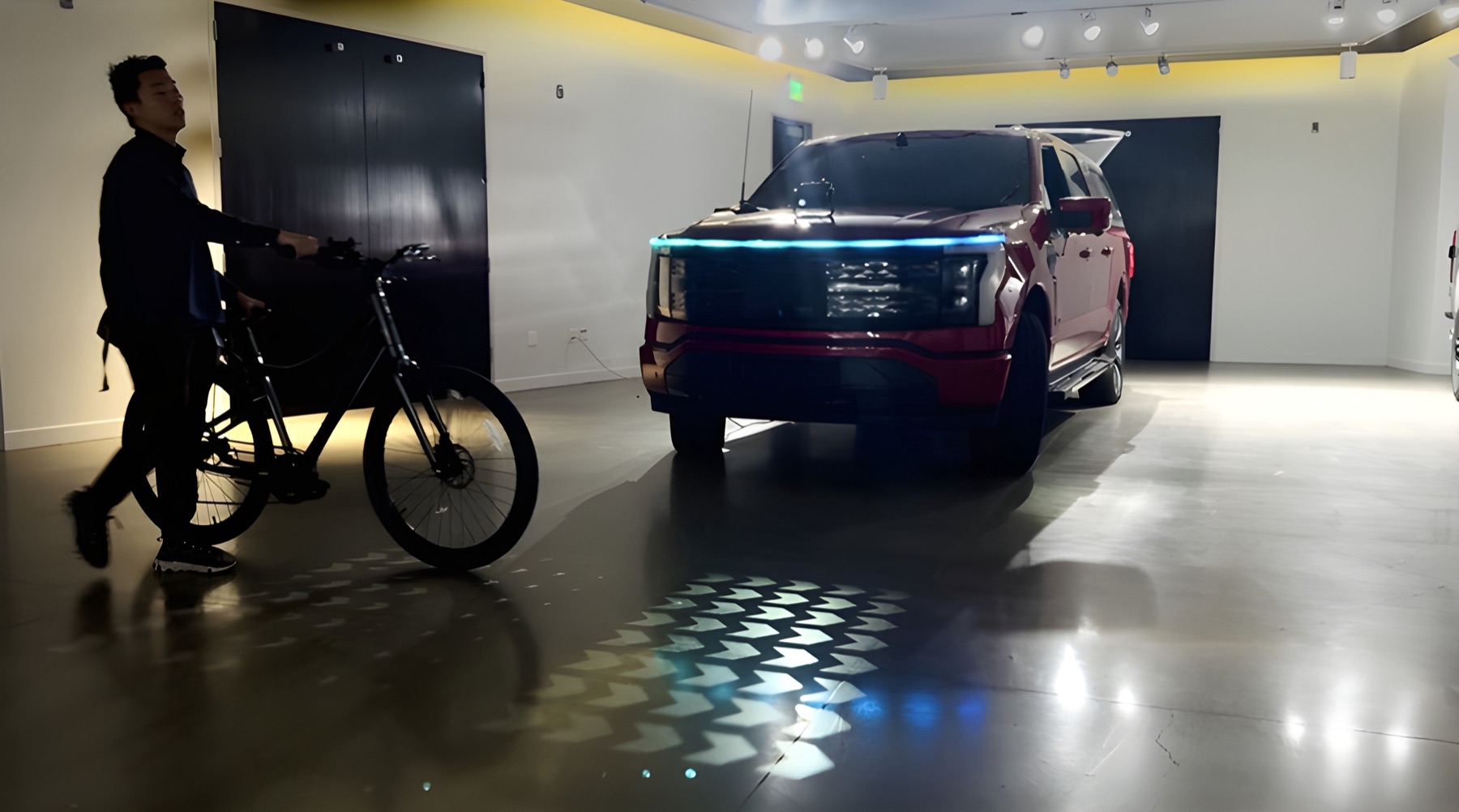 A red electric pickup in a studio, its front light bar lit blue as it projects an animated crosswalk pattern onto the floor toward a person walking a bicycle across its path